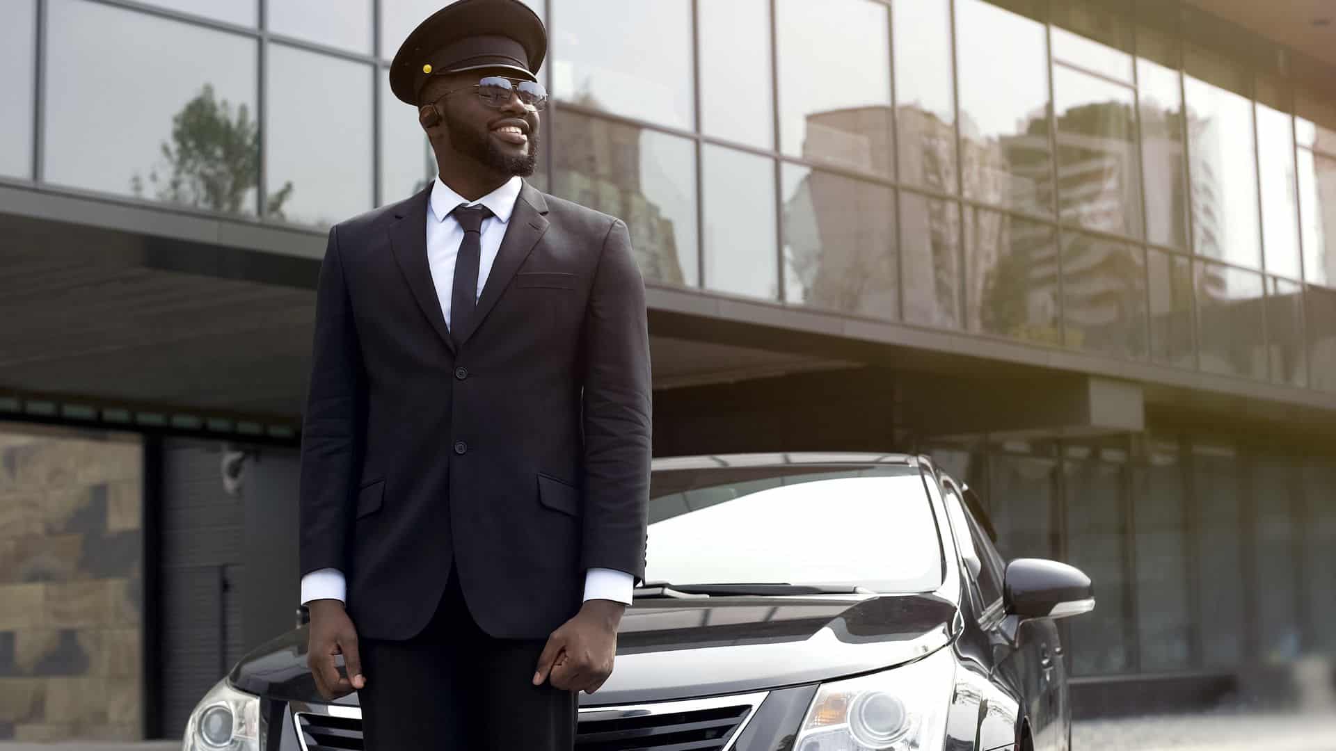 A man in a suit waits for his passengers in front of a private car transfer outside an airport.